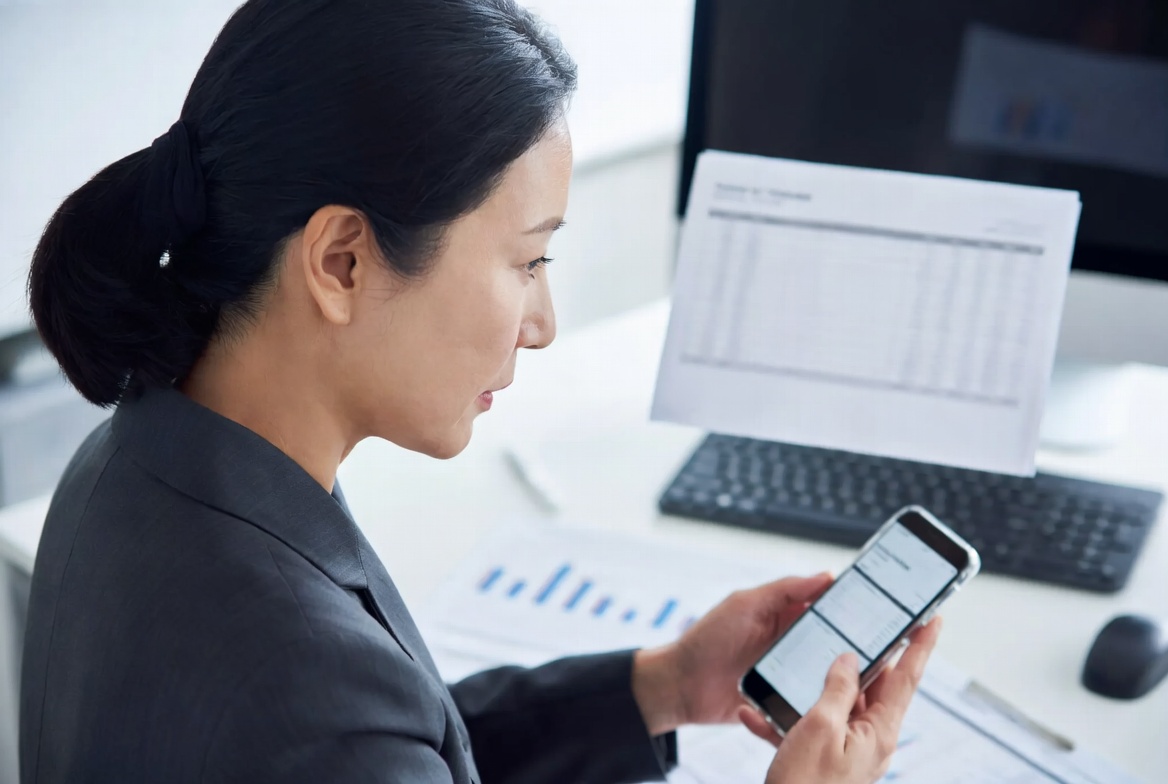 기업 오피스에서 재무 이사가 스마트폰과 장부를 통해 매월 말 상시근로자 수 평균을 모니터링하며 채용 계획을 수립하는 모습